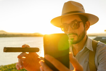 Latino hipster in a lakeside bar smoking a cigar and taking a selfie at sunset