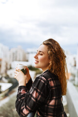 A young, brunette woman dressed in sleepwear enjoys her morning coffee outdoors on the terrace under warm sunlight. City lifestyle . © Алина Битта