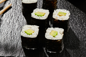 Avocado hosomaki on dark stone table. Hosomaki with avocado. Sushi roll with avocado inside. Style concept japanese menu with black background, leaves and hard shadow.