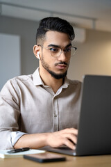 Headshot of young serious indian man working online looking at laptop screen