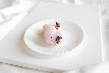 A pink heart-shaped cake lies on a white plate on a light background. Light macaroons on a white canvas. Valentine's Day background
