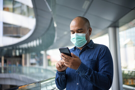 Young indian using smartphone standing in front of office building. Asian man wearing protective mask during pandemic.