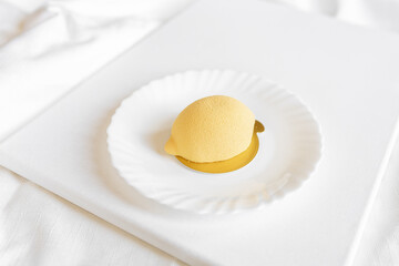 Yellow mousse cake in the form of a lemon lies on a white plate on a light background. Interesting and tasty food. Culinary background