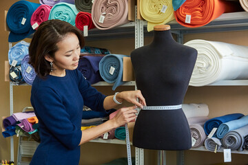 Beautiful Asian woman fashion designer is measuring the waist of a mannequin with a centimeter tape in her workshop