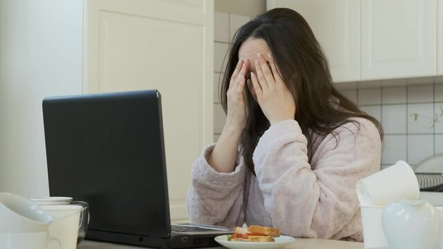 Exhausted Female Suffers from Headache while Using Laptop in the Kitchen. Lots of Empty Coffee Cups Scattered All Over the Table. Working Overtime, Remote Work and Freelancer Lifestyle Concept