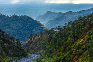 A dry river bed with stones passing through mountains and hills with lush green vegetation and a cloudy sky