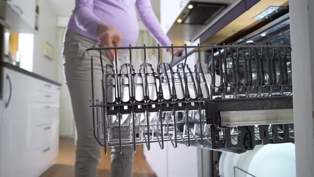 Pregnant Young Woman Taking Out Clean Dishes from a Dishwasher Machine in a Bright Sunny Kitchen. Reliable and Safe Household Appliances for Sustainable Lifestyles. Housewife Uses Modern Appliance to