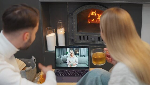 Handheld, young couple sits by the fireplace in living room and communicates with grandmother on a video call using a laptop, holiday time.