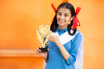 Young beautiful happy indian school girl celebrating victory with trophy,Cheerful braided female holding winning prize against orange background, skill india. child dreams