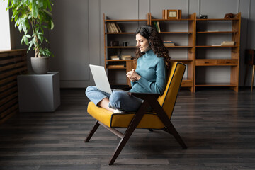 Young woman freelancer sitting with crossed legs on chair at comfortable remote workplace,...