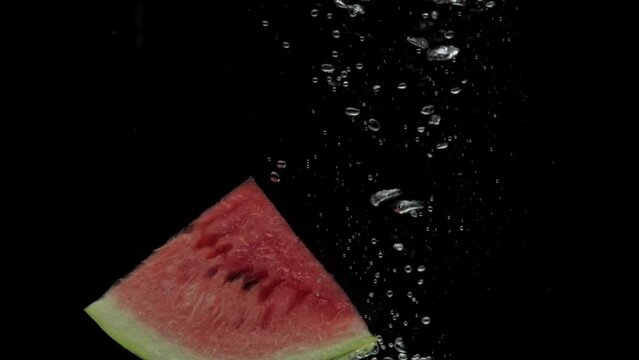 Slow motion slice of watermelon falling into transparent water on black background. Fresh fruit splashing in aquarium. Organic fruit, healthy food, diet, air bubbles