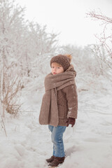 a little handsome boy in knitted winter clothes of brown color walks through a snow-covered winter forest