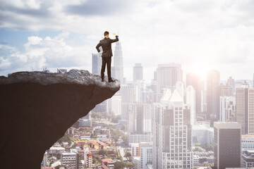 Attractive young european male on cliff edge looking into the distance on bright city background with daylight and mock up place. Future, success, leadership and career growth concept.