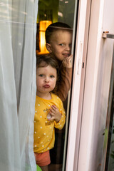 Two small children are standing behind a glass door. The relationship between siblings. The children are locked up at home.