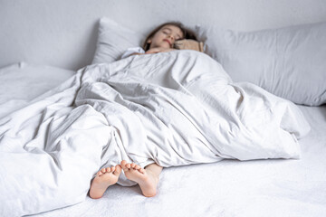 The little girl's legs stick out from under the white blanket.