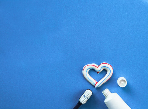 Toothbrush, white tube of toothpaste on blue background. Toothpaste in shape of heart. Top view, copy space. Dental and healthcare concept.