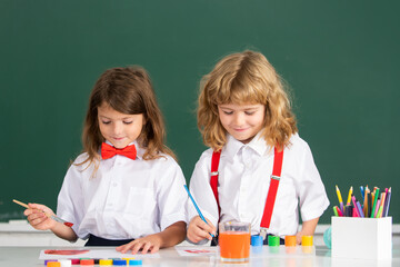 Kids early arts and crafts education. Funny pupils draws in classroom on school blackboard background. School friends kids boy and girl painting together in class.