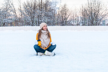 Portrait of happy young woman in yellow sweater and ice skates sitting on the snow, holding mug with coffee and looking away