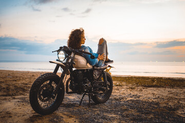 Obraz na płótnie Canvas Beautiful girl having fun driving her custom cafe racer motorcycle, enjoying the sunset on the beach