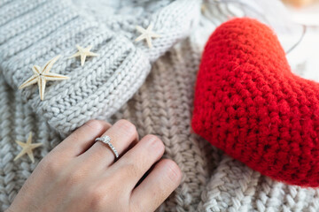Close up of elegant diamond ring on female finger in morning light with red heart shape and sweater background. relationship and love concept. (soft and selective focus)