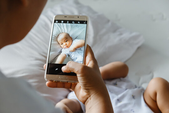 Above view shot of happy mom taking photo her Asian newborn baby boy on bed with smartphone at the home. It is a memorable authentic feeling for every parent when their child is growing up. Top-down