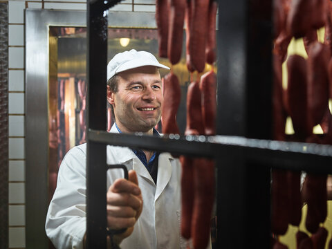 Smiling butcher pulling sausages out of smokehouse at butchery
