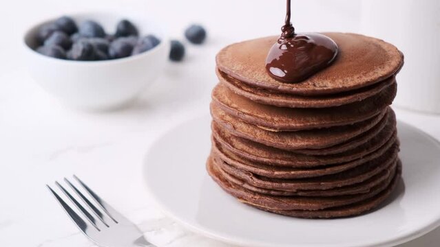 Stack of chocolate pancakes pouring over chocolate sauce, white background.