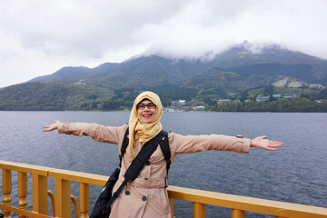 ortrait of smiling muslim woman on board Hakone sightseeing cruise ship at Lake Ashi with mountain fog and cloudy sky background. Waving hands gesture and happy expression.