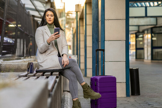 Full length portrait of a smiling successful business woman with a suitcase sitting and talking on the phone in the city.