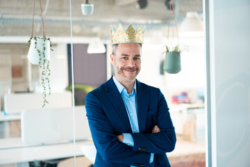 Smiling businessman wearing crown leaning on glass wall with arms crossed at workplace