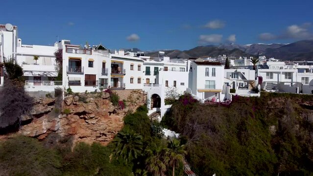 Buildings built on the cliffs surrounding Playa del Carabeo in the city center of Nerja Spain. White-colored stone houses in Nerja city. Blue sky with clouds in the background.