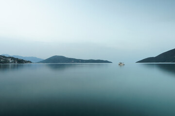 Small ship in the fog on the surface of water with mountains on the background. Bleu and gray tones
