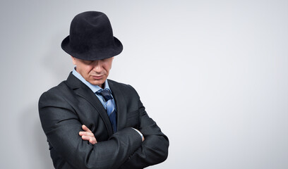 Confident man in a dark suit crossed his arms over his chest and pulled his hat over his eyes, on light gray background. File contains a path to isolation.