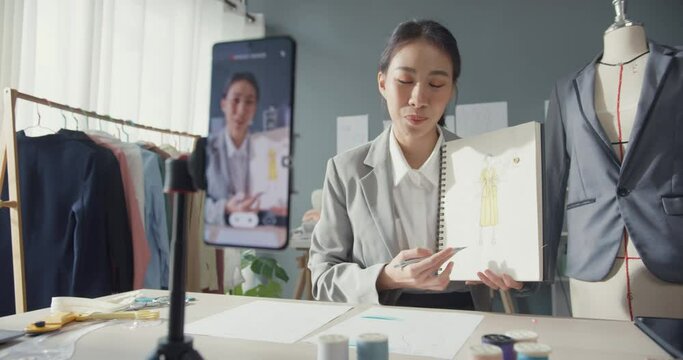 Professional cheerful Asian business woman designer teacher sitting on table look at smartphone online conference share idea tutorial how to made dress in sketchbook to student at studio workplace.