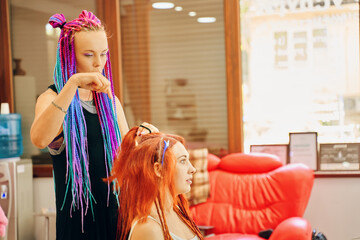 Beautiful girl barber with colored pigtails, who braids ginger dreadlocks to woman. Creative and fashionable hairstyle. Service in beauty salon. Large red chair on background.