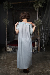 The girl is standing in a pale blue wide dress in a dark room with her back.