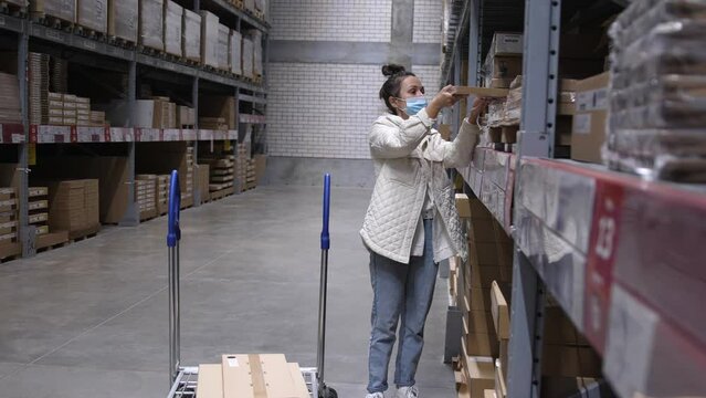 Young woman in a medical mask with shopping cart walking for choosing new furniture in big store warehouse and put some box in a trolley
