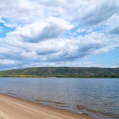 The sandy shore of the big Volga river against the background of blue cloudy sky. On the opposite bank of the Zhiguli Mountains