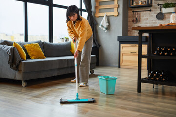 Full length portrait of young Asian woman mopping floors while cleaning cozy apartment