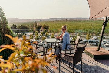 A middle-aged woman sits in a street cafe overlooking the mountains at sunset. She is dressed in a blue jacket and drinks coffee admiring the nature. Travel and vacation concept.