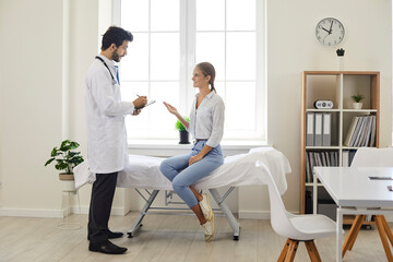 Fototapeta premium Patient talking to a doctor at a modern clinic or hospital. Male physician in a white coat giving a consultation to a young woman who is sitting on the medical couch in the exam room