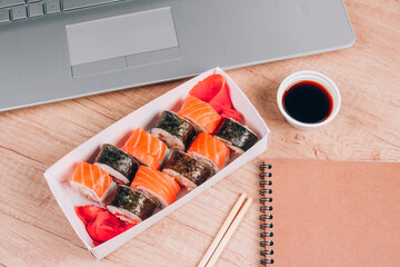 Different sushi rolls with salmon in delivery paper box on wooden table background with laptop. Online home order food and delivery food concept. Takeout food