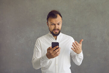 Headshot portrait of confused angry annoyed man using smartphone standing over grey background. Caucasian guy received bad news, looking at broken device gadget with irritation and indignance