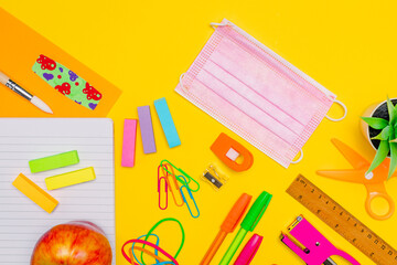 Series of stationery, colored pens, labels, paper clips, calculator and more on yellow background with pink face mask, back to school concept, flat lay