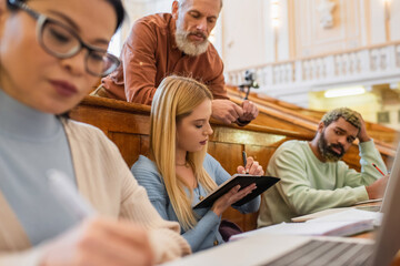 Student writing on notebook near interracial friends and teacher in university