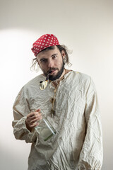 Portrait of a young dark-haired man with a beard, wearing a red cloth bandana on his head and an old shirt with a bottle of rum and a smoking pipe in his teeth on a light background. Pirate Costume,