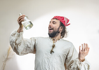 Portrait of a young dark-haired man with a beard with a red cloth bandana on his head and an old shirt with a bottle of rum on a light background. Pirate Costume