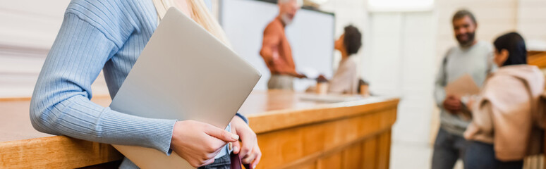 Student holding laptop near blurred friends in university, banner