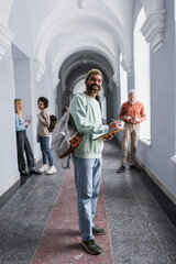 Cheerful african american student with backpack writing on notebook near blurred friends and...