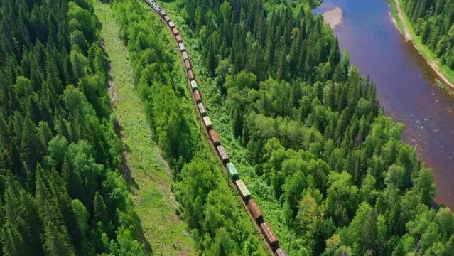Aerial, top view of train moving along river trough forest. Carriages, cars ride on rails on summer day. Railroad transportation of goods taken by drone. Railway cargo, freight delivery services